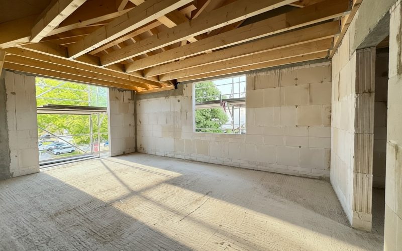 Innenansicht eines Gebäudes im Rohbau mit unverputzten Wänden, sichtbarer Holzdachkonstruktion und großen Fensteröffnungen. Sonnenlicht fällt in den noch unausgebauten Raum.