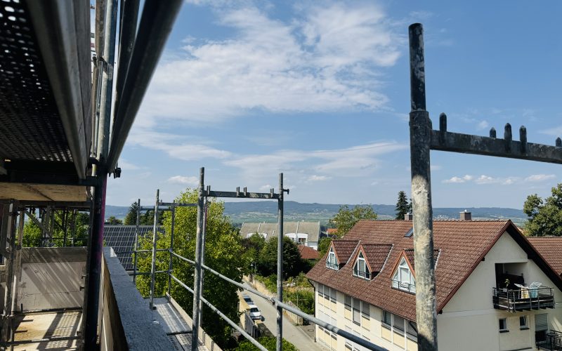 Blick von einem eingerüsteten Gebäude über eine Wohnstraße mit Häusern und Bäumen auf die umliegende Hügellandschaft unter blauem Himmel.