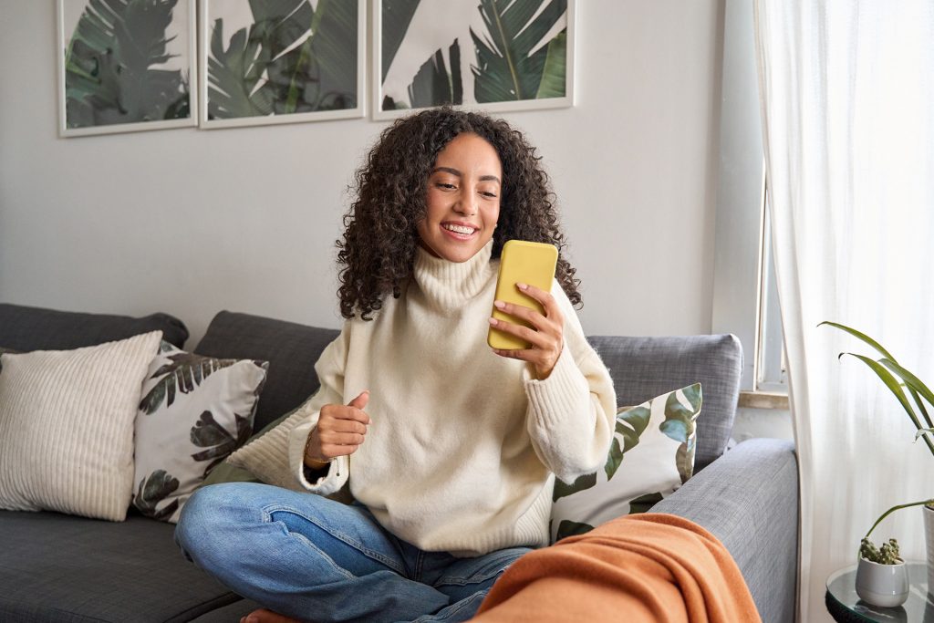 Person mit lockigem Haar sitzt im Schneidersitz auf einem Sofa, hält ein gelbes Smartphone und trägt einen hellen Rollkragenpullover und blaue Jeans.