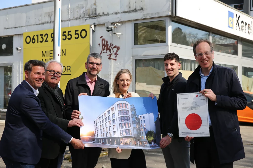 Sechs Personen stehen vor einem Gebäude, halten gemeinsam eine Architekturzeichnung eines modernen mehrstöckigen Gebäudes und ein Dokument mit großem rotem Siegel