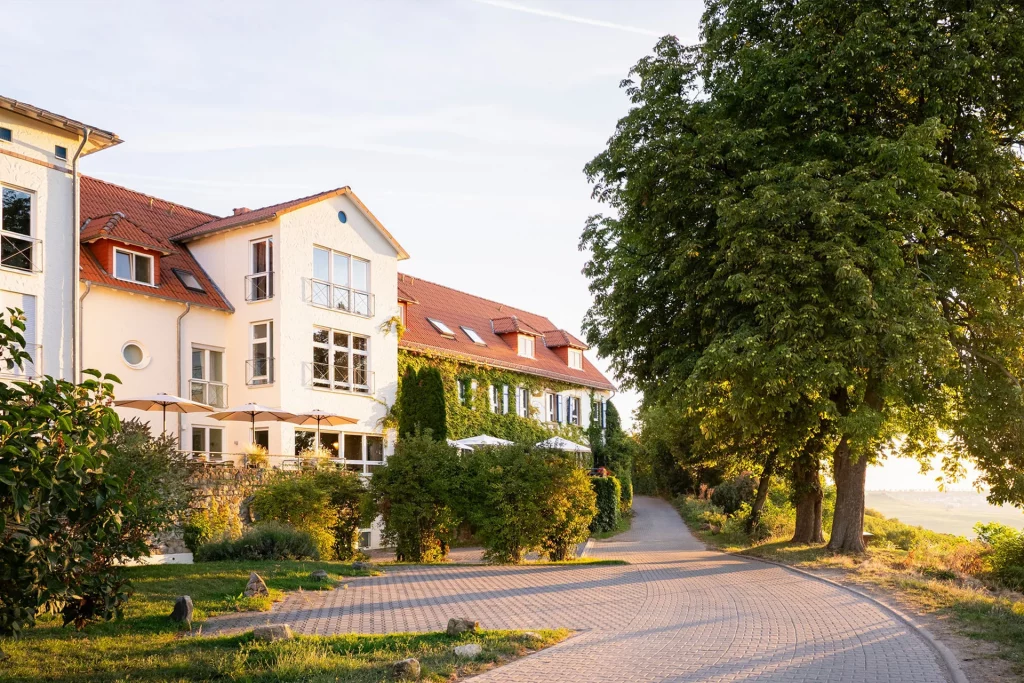 Großes weißes Landhaus mit roten Dachziegeln neben gepflastertem Weg und großen Bäumen bei sonnigem Wetter