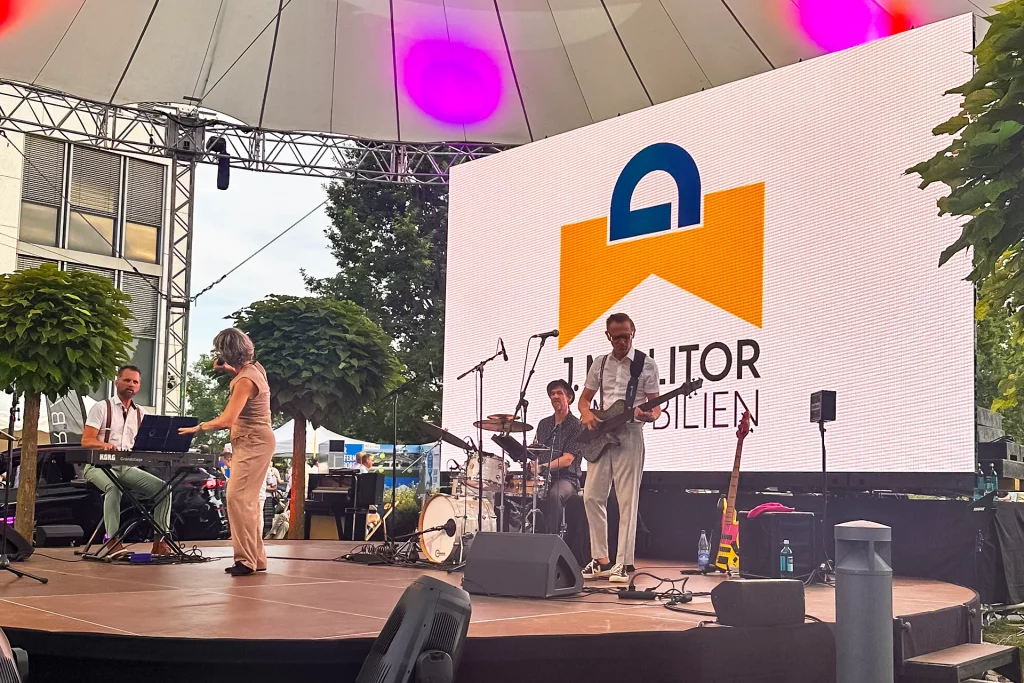 Liveband mit Sängerin, Keyboarder, Schlagzeuger und Gitarrist auf einer überdachten Bühne vor einer großen LED-Wand mit dem Logo von J. Molitor Immobilien.