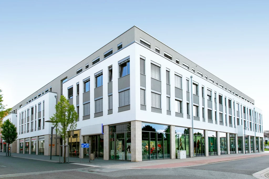Modernes Geschäfts- und Bürogebäude mit weiß-grauer Fassade, großen Fensterflächen und Ladenlokalen im Erdgeschoss an einer Straßenecke.