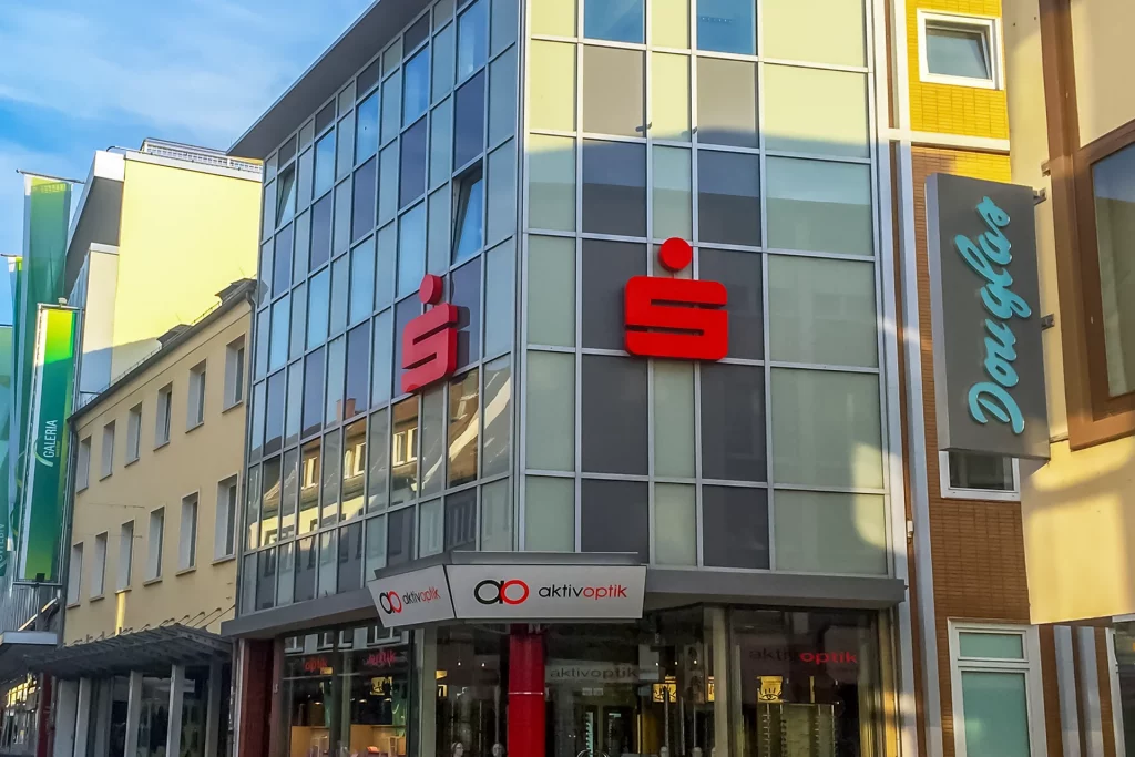 Geschäftshaus mit moderner Glasfassade, roten Sparkassen-Logos und einem Optikergeschäft im Erdgeschoss.