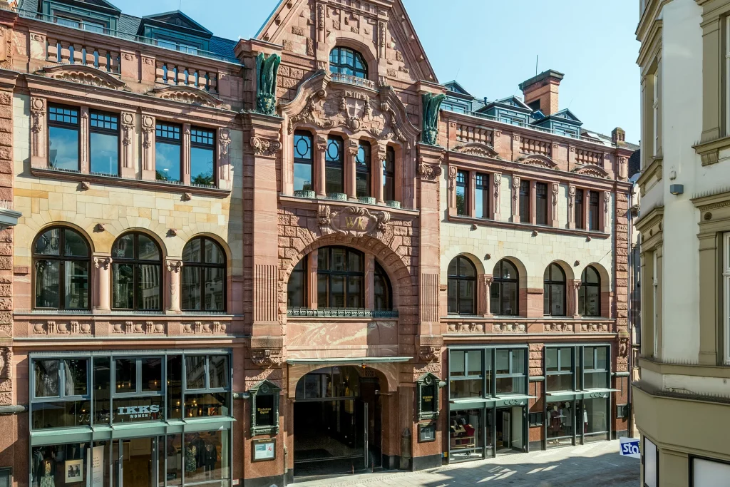 Historisches Geschäftshaus mit reich verzierter Sandsteinfassade, Rundbogenfenstern und zentralem Durchgang. Im Erdgeschoss befinden sich mehrere Ladenlokale.