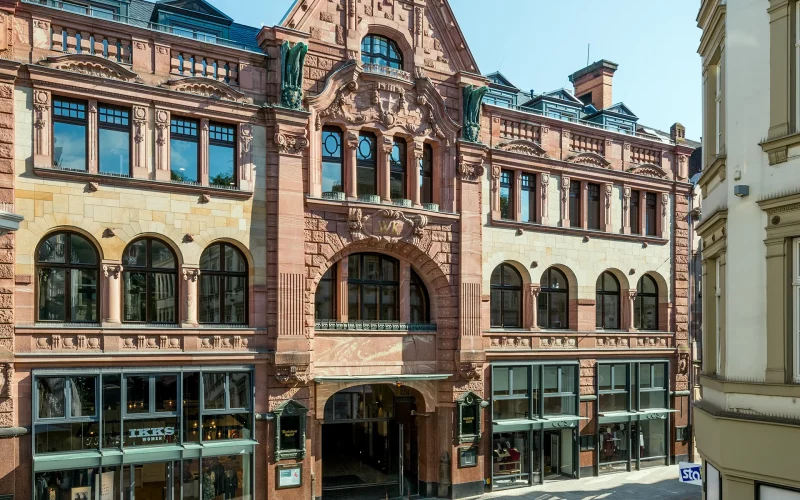 Historisches Geschäftshaus mit reich verzierter Sandsteinfassade, Rundbogenfenstern und zentralem Durchgang. Im Erdgeschoss befinden sich mehrere Ladenlokale.