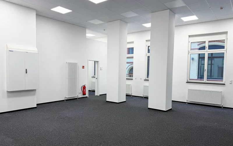 Heller, großzügiger Büroraum mit grauem Teppichboden, weißen Säulen, abgehängter Decke und mehreren Fenstern.