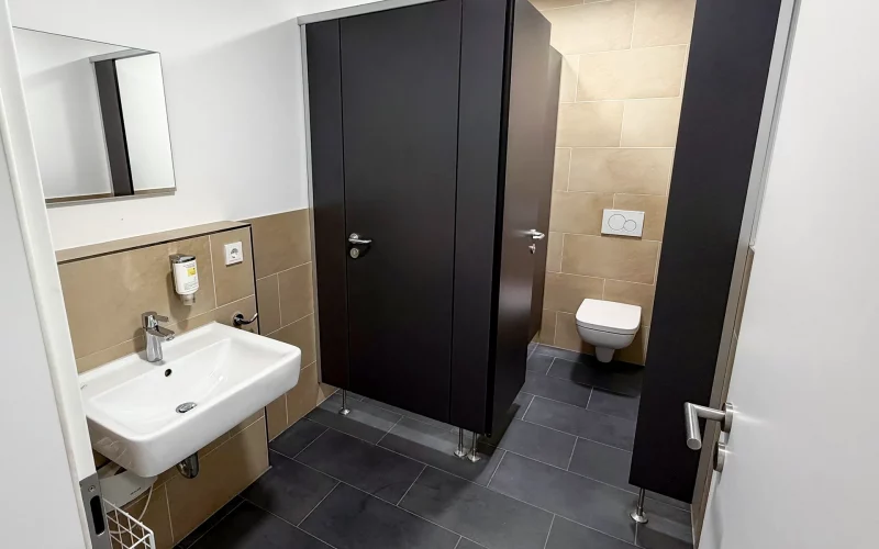 Modern ausgestatteter Sanitärraum mit Waschbecken, Spiegel, dunklen WC-Trennwänden und wandhängender Toilette.