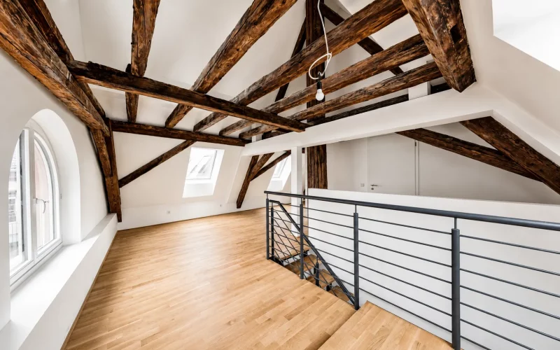 Heller Dachgeschossraum mit Holzboden, markantem Dachgebälk, mehreren Dachfenstern und moderner offener Treppe mit Metallgeländer.