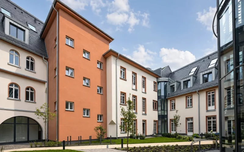 Begrünter Innenhof eines sanierten Gebäudekomplexes mit hellen und terrakottafarbenen Fassaden, historischen Fenstern und modernen Glaselementen.