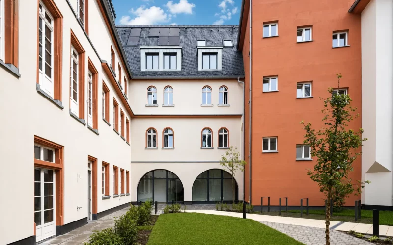 Sanierter Innenhof mit hellen und terrakottafarbenen Fassaden, begrüntem Rasen, jungen Bäumen und historischen Rundbogenfenstern.