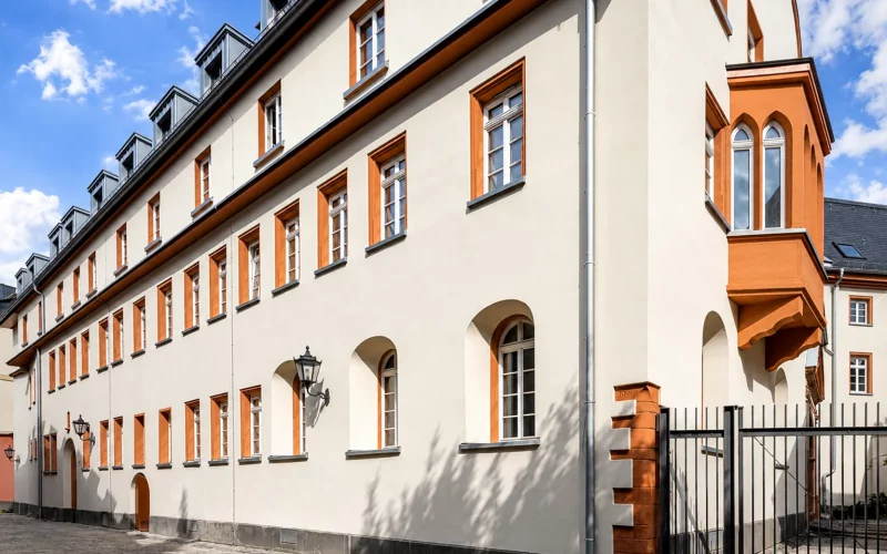 Historisches, langgestrecktes Gebäude mit heller Fassade, orangefarbenen Fensterrahmungen und einem markanten Erker unter blauem Himmel.