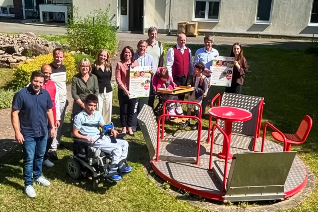 Gruppe von Erwachsenen und Kindern, darunter Personen im Rollstuhl, steht im Freien neben einem barrierefreien roten Karussell. Einige halten Informationsplakate in die Kamera.
